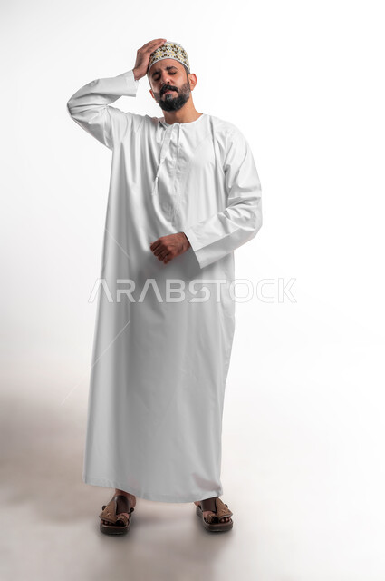 الاحساس بالشقيقية والصداع النصفي, الشعور بالإرهاق وفقدان التركيز, الاجهاد والضغط النفسي, بورتريه لشاب عربي خليجي عماني يرتدي الدشداشة و الكمه يضع يديه على رأسه ويغمض عينيه بإيماءات ألم الرأس الشديد, صورة الجسم بالطول الكامل, خلفية بيضاء