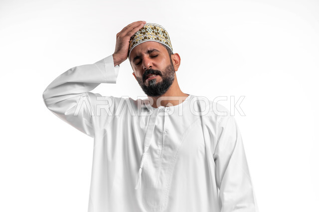 الاحساس بالشقيقية والصداع النصفي, الشعور بالإرهاق وفقدان التركيز, الاجهاد والضغط النفسي, بورتريه مقرب لشاب عربي خليجي عماني يرتدي الدشداشة و الكمه يضع يديه على رأسه ويغمض عينيه بإيماءات ألم الرأس الشديد, خلفية بيضاء