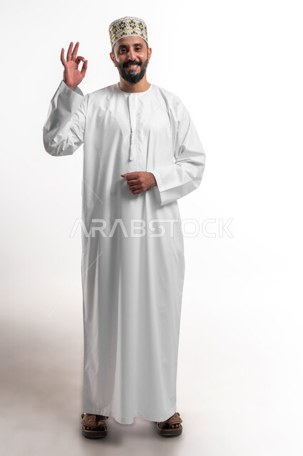 حركات الاعجاب والقبول, بورتريه لشاب عربي خليجي عماني مبتسم يرتدي الدشداشة و الكمه ينظر الى الكاميرا يرفع يديه للأعلى بإيماءات علامة الجودة, تعابير الرضا والسرور والسعادة، صورة الجسم بالطول الكامل، خلفية بيضاء