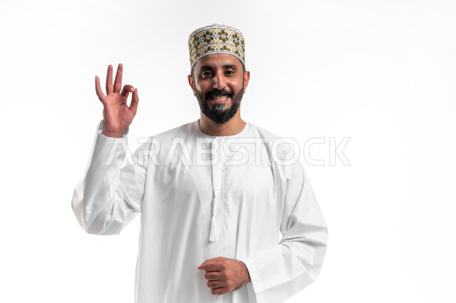 حركات الاعجاب والقبول, بورتريه مقرب لشاب عربي خليجي عماني مبتسم يرتدي الدشداشة و الكمه ينظر الى الكاميرا يرفع يديه للأعلى بإيماءات علامة الجودة, تعابير الرضا والسرور والسعادة، خلفية بيضاء