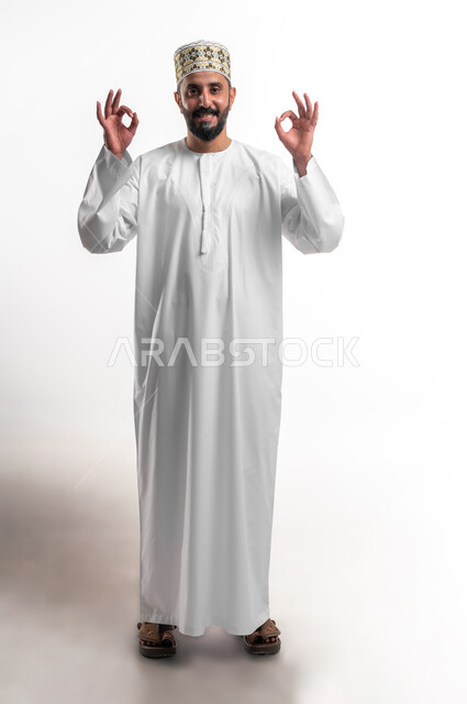 حركات الاعجاب والقبول, بورتريه لشاب عربي خليجي عماني مبتسم يرتدي الدشداشة و الكمه ينظر الى الكاميرا يرفع يديه للأعلى بإيماءات علامة الجودة, تعابير الرضا والسرور والسعادة، صورة الجسم بالطول الكامل، خلفية بيضاء
