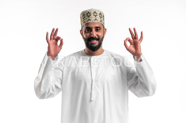 حركات الاعجاب والقبول, بورتريه مقرب لشاب عربي خليجي عماني مبتسم يرتدي الدشداشة و الكمه ينظر الى الكاميرا يرفع يديه للأعلى بإيماءات علامة الجودة, تعابير الرضا والسرور والسعادة، خلفية بيضاء