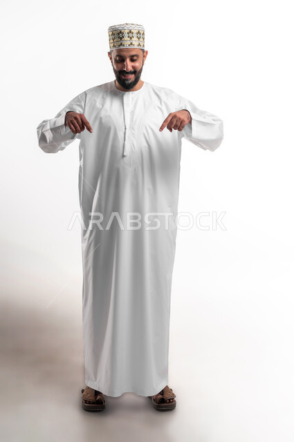 الدهشة والذهول بوجود شيء ما، بورتريه لشاب عربي خليجي عماني مبتسم يرتدي الدشداشة و الكمه يشير بأصابعه نحو الأسفل بإيماءات تدل على هذا المكان (هنا)، صورة الجسم بالطول الكامل، خلفية بيضاء