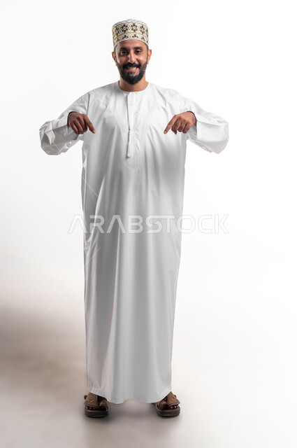 الدهشة والذهول بوجود شيء ما، بورتريه لشاب عربي خليجي عماني مبتسم يرتدي الدشداشة و الكمه ينظر الى الكاميرا يشير بأصابعه نحو الأسفل بإيماءات تدل على هذا المكان (هنا)، صورة الجسم بالطول الكامل، خلفية بيضاء