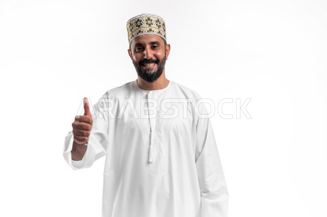 الشعور بالقبول والرضا، بورتريه مقرب لشاب عربي خليجي عماني مبتسم يرتدي الدشداشة و الكمه يرفع اصبع الابهام للاعلى بإيماءات الاعجاب والسرور، خلفية بيضاء