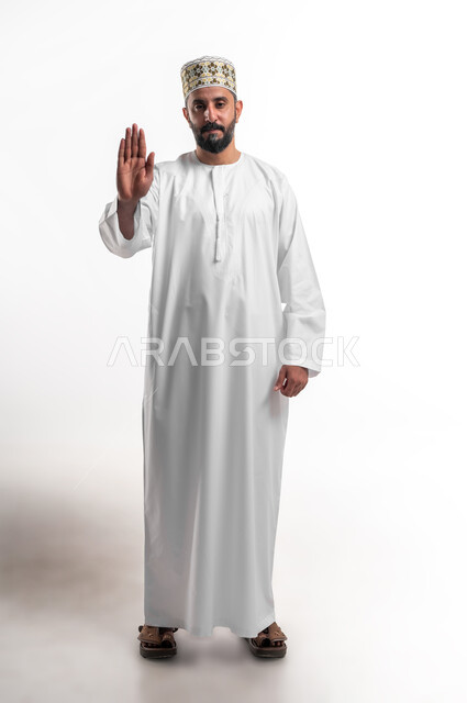 الأمر بالتوقف عن فعل شيء ما، تعابير الرفض والإمتناع، بورتريه لشاب عربي خليجي عماني يرتدي الدشداشة و الكمه يرفع يده بإيماءات النهي، الشعور بالإنزعاج والغضب، صورة الجسم بالطول الكامل، خلفية بيضاء