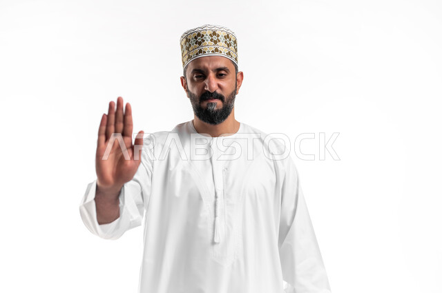 الأمر بالتوقف عن فعل شيء ما، تعابير الرفض والإمتناع، بورتريه مقرب لشاب عربي خليجي عماني يرتدي الدشداشة و الكمه يرفع يده بإيماءات النهي، الشعور بالإنزعاج والغضب، خلفية بيضاء