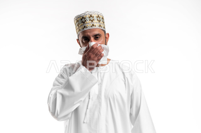 المرض والرشح والكحه والانفلونزا, الإحساس بالكسل والخمول، بورتريه مقرب لشاب عربي خليجي عماني يرتدي الدشداشة و الكمه يمسك بيده منديل، الاصابة بفايروس كورونا المستجد، صورة الجسم بالطول الكامل، خلفية بيضاء