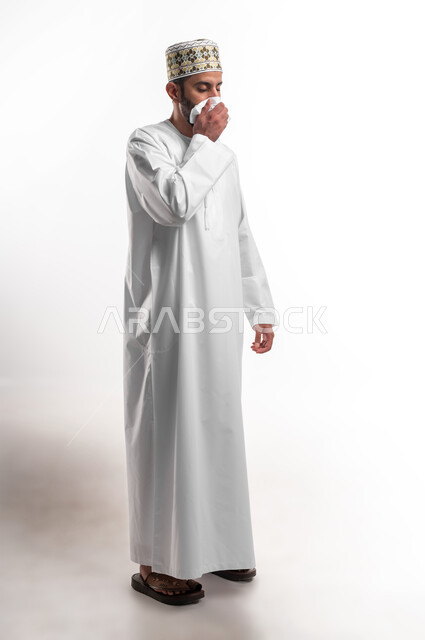 المرض والرشح والكحه والانفلونزا, الإحساس بالكسل والخمول، بورتريه لشاب عربي خليجي عماني يرتدي الدشداشة و الكمه يمسك بيده منديل، الاصابة بفايروس كورونا المستجد، صورة الجسم بالطول الكامل، خلفية بيضاء