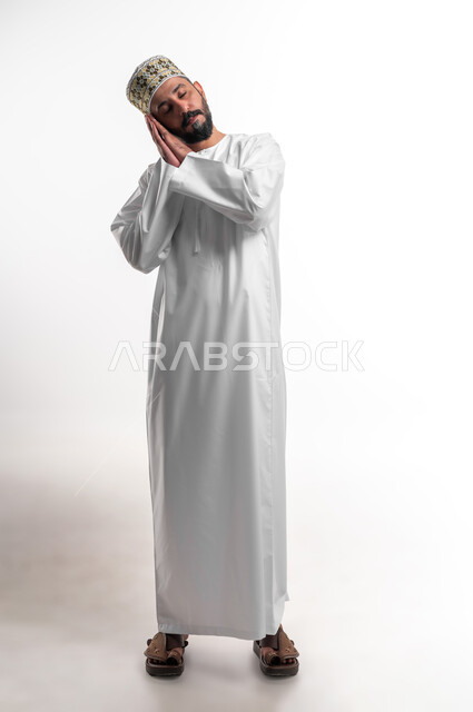 الشعور بالتعب و الحاجه الى الراحه, الإحساس بالكسل والخمول، بورتريه مقرب لشاب عربي خليجي عماني يرتدي الدشداشة و الكمه يضع يديه تحت رأسه يغمض عينيه بإيماءات النوم، تعابير النعاس والإرهاق، صورة الجسم بالطول الكامل، خلفية بيضاء