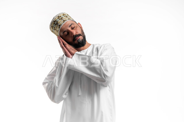 الشعور بالتعب و الحاجه الى الراحه, الإحساس بالكسل والخمول، بورتريه مقرب لشاب عربي خليجي عماني يرتدي الدشداشة و الكمه يضع يديه تحت رأسه يغمض عينيه بإيماءات النوم، تعابير النعاس والإرهاق، خلفية بيضاء