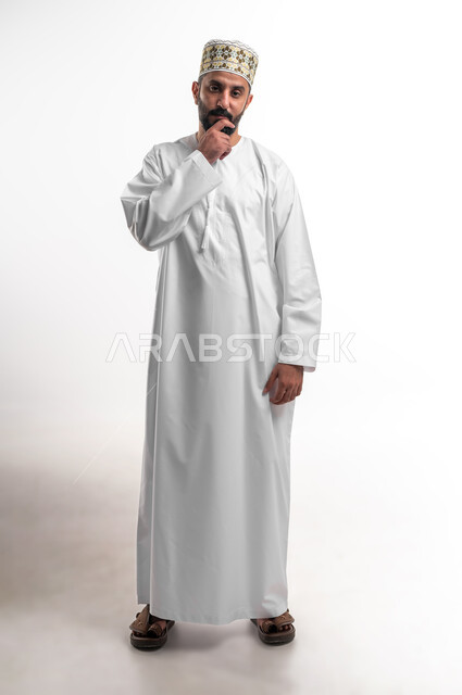 Contemplation and concentration to find the solution to a problem, feeling upset and bored, solving problems and making decisions, portrait of a young Arab Gulf Omani man wearing a dishdasha and a sleeve, placing his hand on his face with gestures of thought, confusion and annoyance, full-length body portrait, white background