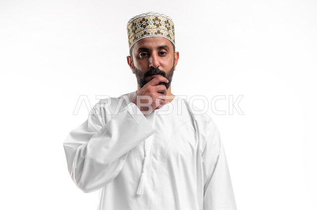 التمعن والتركيز لإيجاد الحل لمشكلة ما, الشعور بالضيق والملل، حل المشكلات واتخاذ القرارات، بورتريه مقرب لشاب عربي خليجي عماني يرتدي الدشداشة و الكمه يضع يده على وجهه بإيماءات التفكير والحيرة والانزعاج، خلفية بيضاء