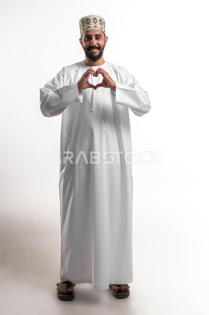 اظهار مشاعر الحب والشغف، مفهوم المحبة والمودة، بورتريه لشاب عربي خليجي عماني يرتدي الدشداشة و الكمه يرفع يديه بإيماءات على شكل قلب، تعابير الفرح والسرور والسعادة، صورة الجسم بالطول الكامل, خلفية بيضاء