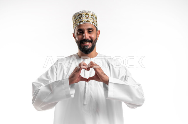 اظهار مشاعر الحب والشغف، مفهوم المحبة والمودة، بورتريه مقرب لشاب عربي خليجي عماني يرتدي الدشداشة و الكمه يرفع يديه بإيماءات على شكل قلب، تعابير الفرح والسرور والسعادة، خلفية بيضاء