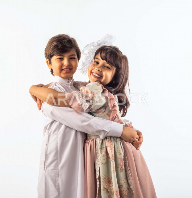 خلفية بيضاء لشقيق وأخته صغيران سعوديان مبتسمان ، يعانقان بعضهما , ينظران إلى الكاميرا , بإيماءات وجه ويدين مختلفة تدل على الحب والأمان , بورتريه لطفلان سعوديان خليجيان أنيقان يرتديان لباس العيد الجميل , الاحتفال بعيد الفطر السعيد , عيد الفطر المبارك