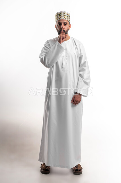 التوقف عن الكلام و الأمر بالصمت والسكوت، بورتريه لشاب عربي خليجي عماني يرتدي الدشداشة و الكمه ينظر الى الكاميرا  يرفع اصبعه الى فمه بإيماءات طلب الهدوء وعدم الازعاج, صورة الجسم بالطول الكامل، خلفية بيضاء