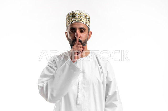 التوقف عن الكلام و الأمر بالصمت والسكوت، بورتريه مقرب لشاب عربي خليجي عماني يرتدي الدشداشة و الكمه ينظر الى الكاميرا  يرفع اصبعه الى فمه بإيماءات طلب الهدوء وعدم الازعاج, خلفية بيضاء