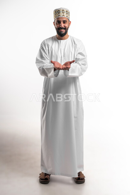 مفهوم التقديم والعرض، بورتريه لشاب عربي خليجي عماني مبتسم يرتدي الدشداشة و الكمه ينظر الى الكاميرا  يفتح كفيه للأعلى بإيماءات حمل عنصر ما, صورة الجسم بالطول الكامل، خلفية بيضاء