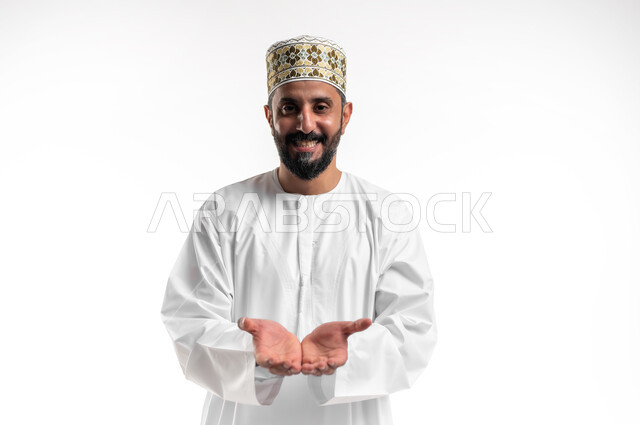 Concept of presentation and presentation, portrait of a smiling Arab Gulf Omani young man wearing a dishdasha and cuff, looking at the camera, opening his palms upwards with gestures of holding an item, white background