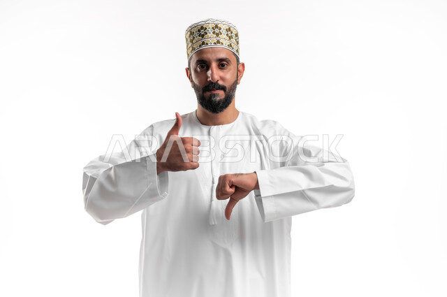 الحيرة والتردد في شيء ما، بورتريه مقرب لشاب عربي خليجي عماني مبتسم يرتدي الدشداشة و الكمه ينظر الى الكاميرا يرفع اصبع الابهام للاعلى والاسفل بإيماءات عدم الموافقه والموافقه عدم الإعجاب والإعجاب, مفهوم الرفض والقبول، خلفية بيضاء