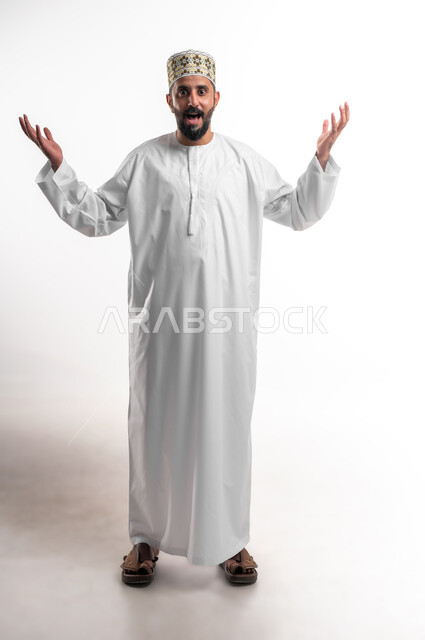 الانتصار والفوز والنجاح، مفهوم الانجاز وتحقيق الاهداف، بورتريه مقرب لشاب عربي خليجي عماني مبتسم يرتدي الدشداشة و الكمه ينظر الى الكاميرا يرفع يديه للاعلى بإيماءات الفرح والسرور, الشعور بالسعادة والقوة، صورة الجسم بالطول الكامل, خلفية بيضاء