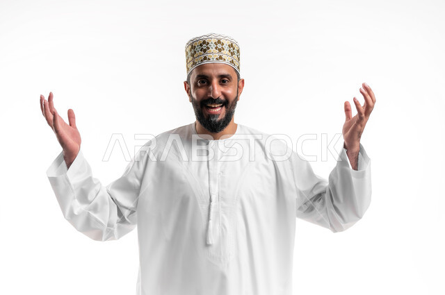 الانتصار والفوز والنجاح، مفهوم الانجاز وتحقيق الاهداف، بورتريه مقرب لشاب عربي خليجي عماني مبتسم يرتدي الدشداشة و الكمه ينظر الى الكاميرا يرفع يديه للاعلى بإيماءات الفرح والسرور, الشعور بالسعادة والقوة، خلفية بيضاء