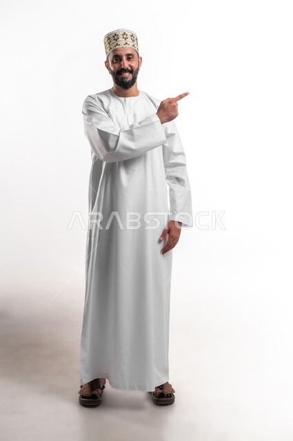 تعابير التأكد والتحديد, بورتريه لشاب عربي خليجي عماني يرتدي الدشداشة و الكمه ينظر الى الكاميرا يشير بأصابع السبابة الى شيء ما بإيماءات السرور، تعابير البهجة والفرح، صورة الجسم بالطول الكامل، خلفية بيضاء