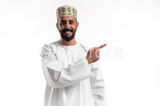 تعابير التأكد والتحديد, بورتريه مقرب لشاب عربي خليجي عماني يرتدي الدشداشة و الكمه ينظر الى الكاميرا يشير بأصابع السبابة الى شيء ما بإيماءات السرور، تعابير البهجة والفرح، خلفية بيضاء