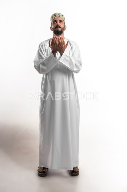 الإكثار من الدعاء في شهر رمضان المبارك، الإجتهاد في العبادة والحفاظ على الطاعات، بورتريه مقرب لشاب عربي خليجي عماني يرتدي الدشداشة و الكمه يرفع يديه ويدعو الله بإيماءات التوسل والتضرع، صورة الجسم بالطول الكامل, خلفية بيضاء