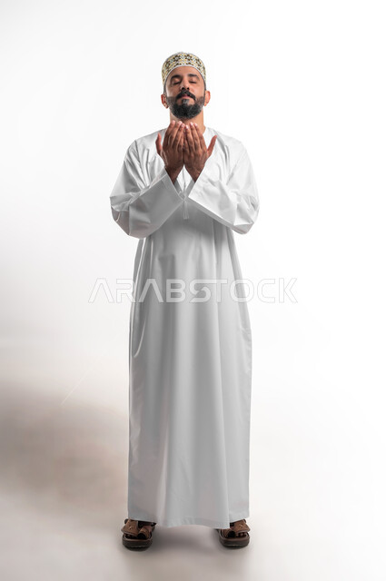 Diligence in worship and maintaining obedience, frequent supplication in the holy month of Ramadan, portrait of a young Arab Gulf Omani man wearing a dishdasha and a sleeve, raising his hands and calling upon God with gestures of supplication and supplication, full-length body portrait, white background