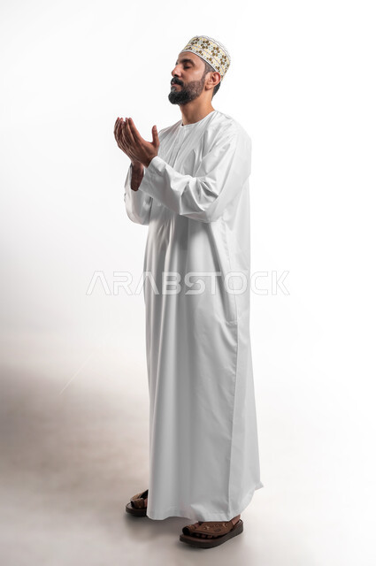Diligence in worship and maintaining obedience, frequent supplication in the holy month of Ramadan, portrait of a young Arab Gulf Omani man wearing a dishdasha and a sleeve, raising his hands and calling upon God with gestures of supplication and supplication, full-length body portrait, white background