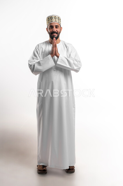 مفهوم الاستقبال والترحيب، الإكثار من الدعاء في شهر رمضان، الإجتهاد في العبادة والحفاظ على الطاعات، بورتريه لشاب عربي خليجي عماني مبتسم يرتدي الدشداشة و الكمه ينظر الى الكاميرا يرفع يديه ويدعو الله بإيماءات التوسل والتضرع، صورة الجسم بالطول الكامل, خلفية بيضاء