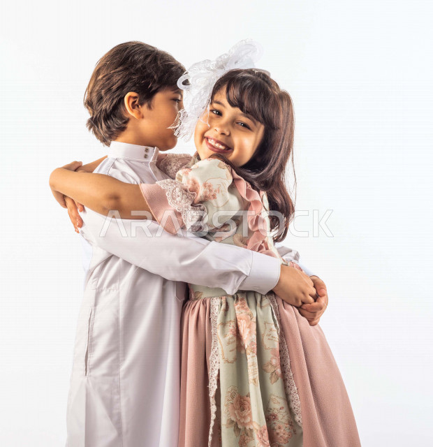 خلفية بيضاء لشقيق وأخته صغيران سعوديان مبتسمان ، يعانقان بعضهما , وينظر الأخ لإخته الصغيرة , بإيماءات وجه ويدين مختلفة تدل على الحب والأمان , بورتريه لطفلان سعوديان خليجيان أنيقان يرتديان لباس العيد الجميل , الاحتفال بعيد الفطر السعيد , عيد الفطر المبارك