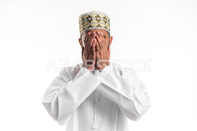 الإكثار من الدعاء في شهر رمضان المبارك، الإجتهاد في العبادة والحفاظ على الطاعات، بورتريه مقرب لشاب عربي خليجي عماني يرتدي الدشداشة و الكمه يضع يديه على وجهه ويدعو الله بإيماءات التوسل والتضرع، خلفية بيضاء