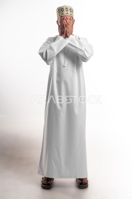 الإكثار من الدعاء في شهر رمضان المبارك، الإجتهاد في العبادة والحفاظ على الطاعات، بورتريه لشاب عربي خليجي عماني يرتدي الدشداشة و الكمه يضع يديه على وجهه ويدعو الله بإيماءات التوسل والتضرع، صورة الجسم بالطول الكامل, خلفية بيضاء