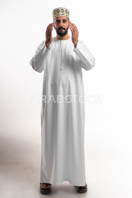 الإكثار من الدعاء في شهر رمضان المبارك، الإجتهاد في العبادة والحفاظ على الطاعات، بورتريه لشاب عربي خليجي عماني يرتدي الدشداشة و الكمه يرفع يديه ويدعو الله بإيماءات التوسل والتضرع، صورة الجسم بالطول الكامل, خلفية بيضاء