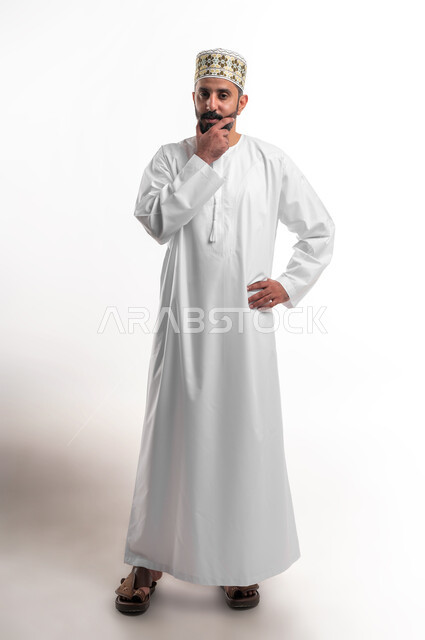 Feeling upset and bored, contemplating and concentrating to find the solution to a problem, solving problems and making decisions, portrait of a young Arab Gulf Omani man wearing a dishdasha and a sleeve, putting his hand on his face with gestures of thought, confusion and annoyance, full-length body portrait, white background