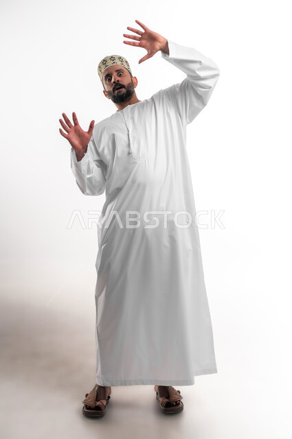 القلق والاحتياط والابتعاد عن شيء ما, حماية النفس من ضرر خارجي, بورتريه لشاب عربي خليجي عماني يرتدي الدشداشة و الكمه يرفع يديه للاعلى بإيماءات التوقف وعدم الاقتراب، مفهوم الحذر والصدمة، صورة الجسم بالطول الكامل, خلفية بيضاء
