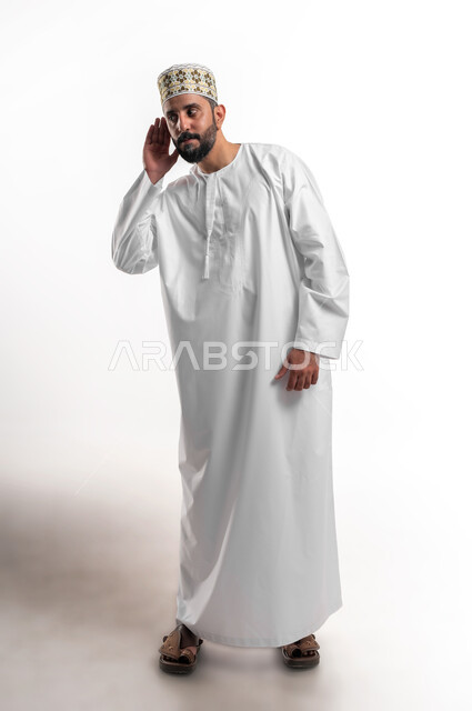 الانصات وحسن الاستماع، التفاجؤ من شيء ما، بورتريه لشاب عربي خليجي عماني مبتسم يرتدي الدشداشة و الكمه يضع يده خلف أذنه بإيماءات التنصت والتجسس، التركيز على الكلام وسماع الاسرار، خلفية بيضاء