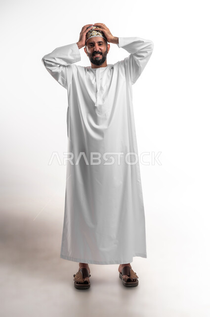 التفاجؤ من شيء ما، الشعور بالدهشة والصدمة والذهول, بورتريه لشاب عربي خليجي عماني مبتسم يرتدي الدشداشة و الكمه ينظر الى الكاميرا ينظر الى الكاميرا يضع يديه على رأسه بإيماءات التعجب والفرح, الشعور بالسعادة والقوة، صورة الجسم بالطول الكامل, خلفية بيضاء