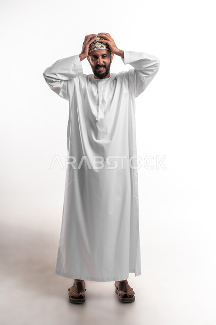 التفاجؤ من شيء ما، الشعور بالدهشة والصدمة والذهول, بورتريه لشاب عربي خليجي عماني مبتسم يرتدي الدشداشة و الكمه ينظر الى الكاميرا ينظر الى الكاميرا يضع يديه على رأسه بإيماءات التعجب والفرح, الشعور بالسعادة والقوة، صورة الجسم بالطول الكامل, خلفية بيضاء