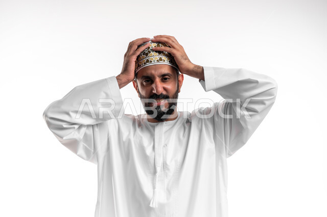 التفاجؤ من شيء ما، الشعور بالدهشة والصدمة والذهول, بورتريه مقرب لشاب عربي خليجي عماني مبتسم يرتدي الدشداشة و الكمه ينظر الى الكاميرا ينظر الى الكاميرا يضع يديه على رأسه بإيماءات التعجب والفرح, الشعور بالسعادة والقوة، خلفية بيضاء