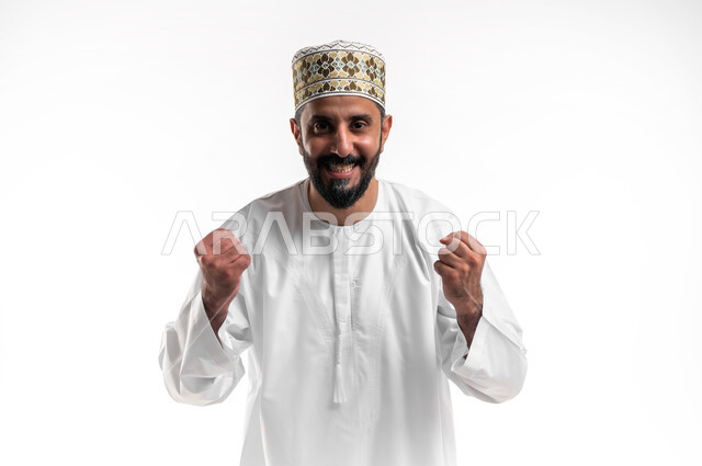 الانتصار والفوز والنجاح، مفهوم الانجاز وتحقيق الاهداف، بورتريه مقرب لشاب عربي خليجي عماني مبتسم يرتدي الدشداشة و الكمه ينظر الى الكاميرا يرفع قبضة يديه للاعلى بإيماءات الفرح والسرور, الشعور بالسعادة والقوة، خلفية بيضاء