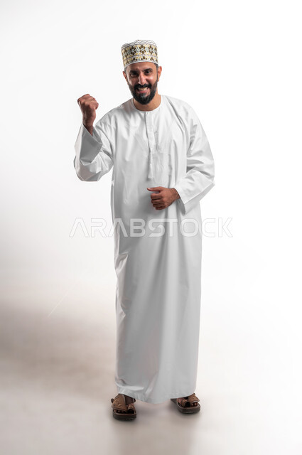 مفهوم الانجاز وتحقيق الاهداف، بورتريه لشاب عربي خليجي عماني مبتسم يرتدي الدشداشة و الكمه ينظر الى الكاميرا يرفع قبضة يده للاعلى بإيماءات الفرح والسرور, الشعور بالسعادة والقوة، الانتصار والفوز والنجاح، صورة الجسم بالطول الكامل, خلفية بيضاء
