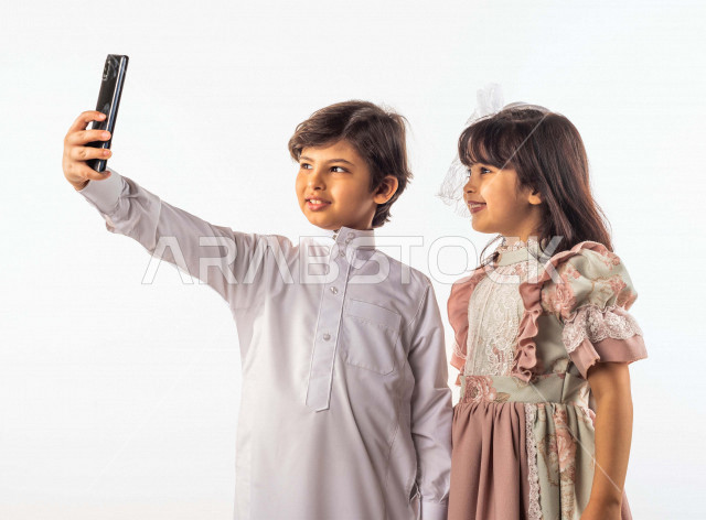 خلفية بيضاء لصبي وفتاه صغيران سعوديان مبتسمان ، يمسك الصبي الجوال بيده , يقومان بإلتقاط صور سيلفي تذكاريه بإستخدام الهاتف المحمول بإيماءات وجه ويدين مختلفة , بورتريه لطفل وطفلة سعوديان خليجيان سعيدان يرتديان لباس العيد ,الاحتفال بعيد الفطر السعيد