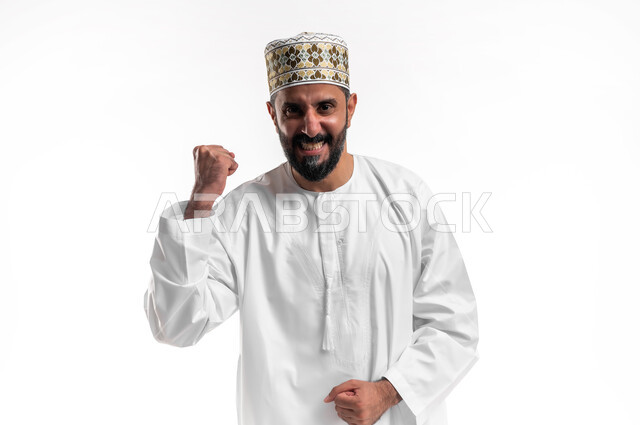 مفهوم الانجاز وتحقيق الاهداف، بورتريه مقرب لشاب عربي خليجي عماني مبتسم يرتدي الدشداشة و الكمه ينظر الى الكاميرا يرفع قبضة يده للاعلى بإيماءات الفرح والسرور, الشعور بالسعادة والقوة، الانتصار والفوز والنجاح، خلفية بيضاء