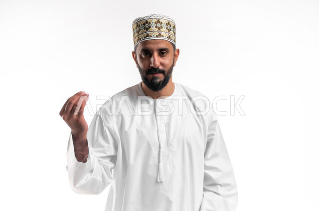 مفهوم الصبر والتريث و التأني, التهديد و التوعد لشيء ما، بورتريه مقرب لشاب عربي خليجي عماني مبتسم يرتدي الدشداشة و الكمه ينظر الى الكاميرا يرفع يده نحو الأعلى بإيماءات طلب الإنتظار, إمهال بعض الوقت لفعل شيء، خلفية بيضاء