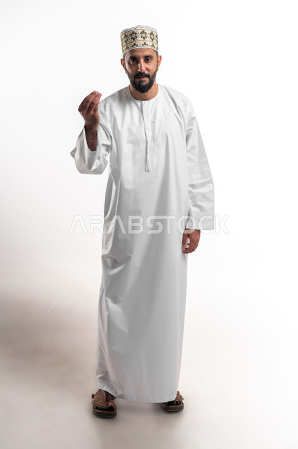 مفهوم الصبر والتريث و التأني, التهديد و التوعد لشيء ما، بورتريه مقرب لشاب عربي خليجي عماني مبتسم يرتدي الدشداشة و الكمه ينظر الى الكاميرا يرفع يده نحو الأعلى بإيماءات طلب الإنتظار, إمهال بعض الوقت لفعل شيء، صورة الجسم بالطول الكامل, خلفية بيضاء