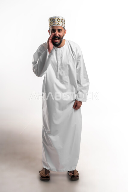 الكلام والنداء والتحدث بصوت مرتفع، بورتريه لشاب عربي خليجي عماني مبتسم يرتدي الدشداشة و الكمه ينظر الى الكاميرا يضع يده على فمه بإيماءات المناداة لشخص ما، دردشة ومحادثات وتواصل اجتماعي، صورة الجسم بالطول الكامل, خلفية بيضاء